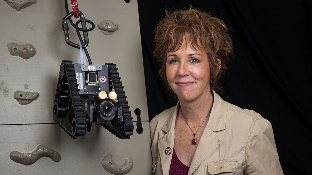 Image of Dr. Robin Murphy posing next to a robot that is hanging from a piece of rock-climbing wall.