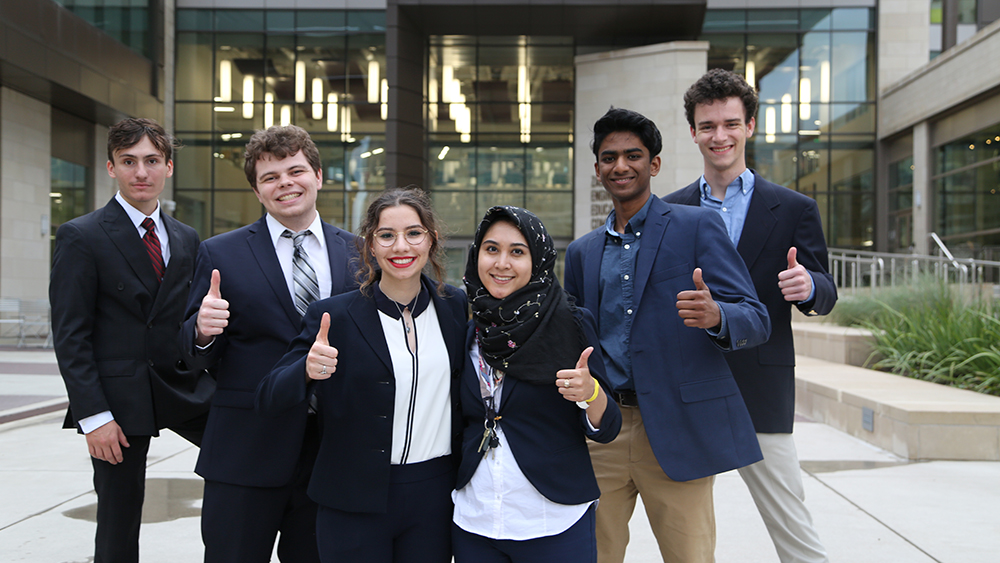 Six students on winning team smile, pose in front of ZACH building with thumbs up.