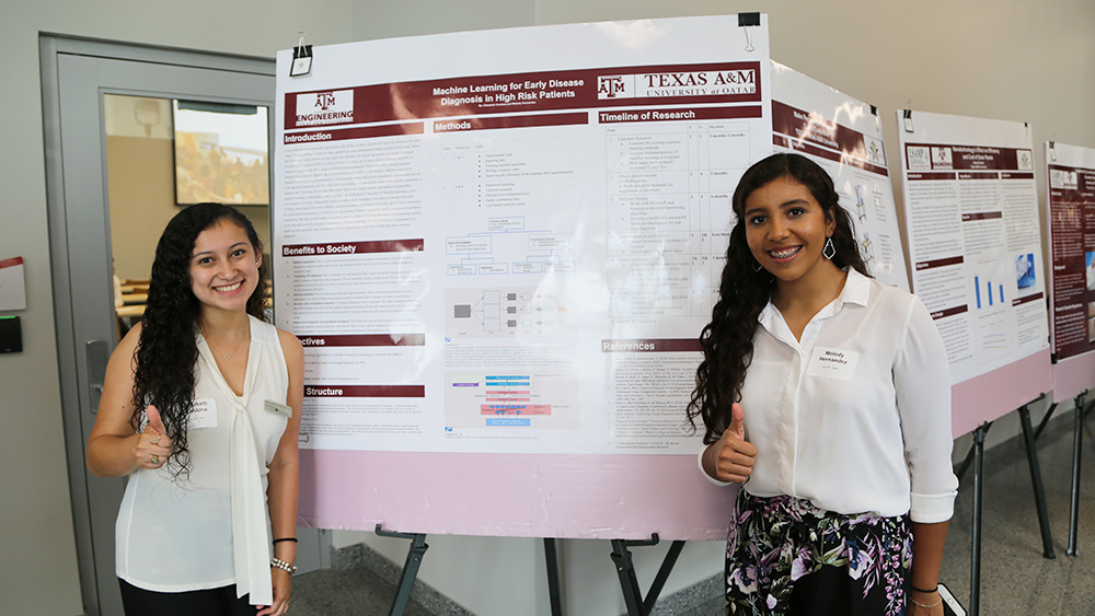 Two female students stand in front of a research poster with their thumbs up