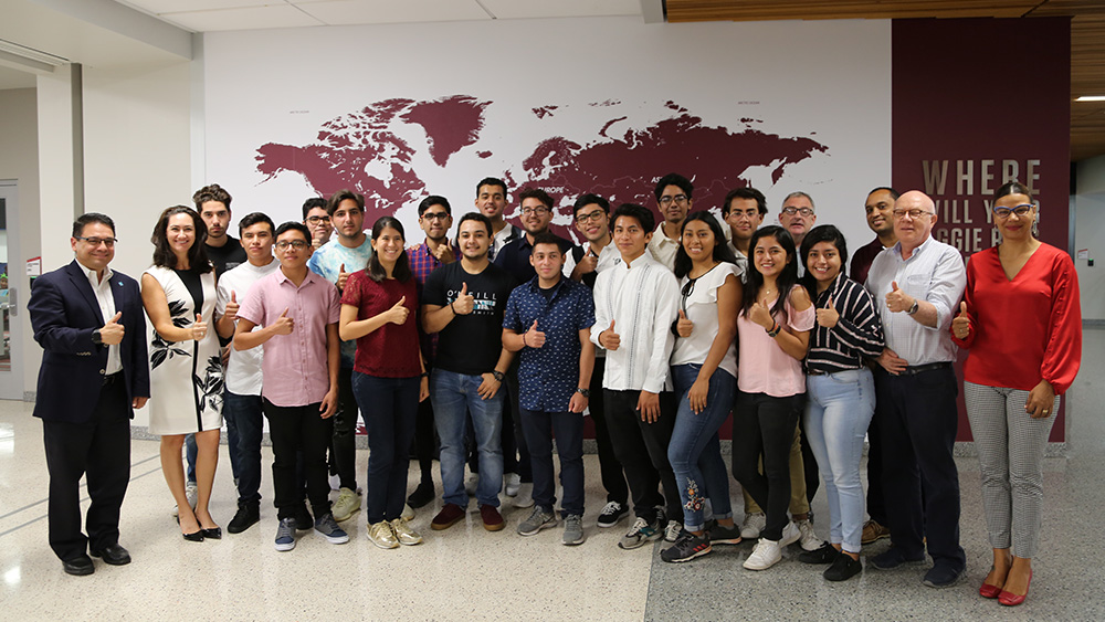 A large group of students and faculty stand in front of a world map on the wall