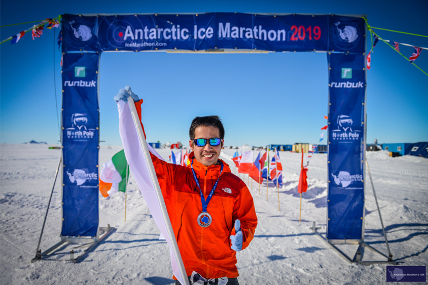 Kentaro Iio standing in front of the finish line at the Antarctic Ice Marathon. White snow and internal flags surround him. 