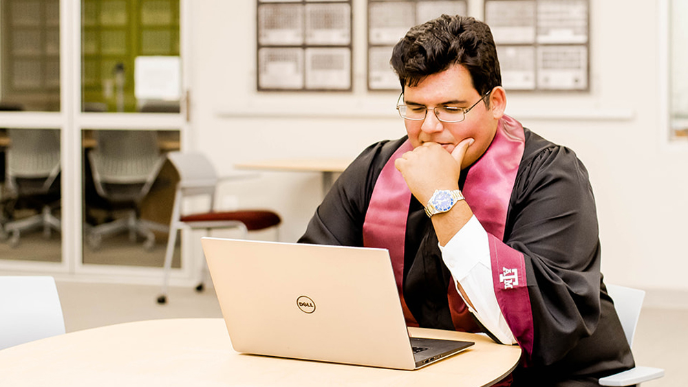 Civil Engineering graduate Andrew Salazar at a computer wearing his graduation gown and stole 
