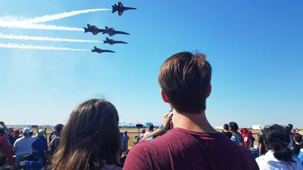 Katie and Steven, former aerospace engineering students, have their backs to the camera. Steven’s arm is around Katie as they look up at five blue and yellow jet planes flying above them at an airshow.