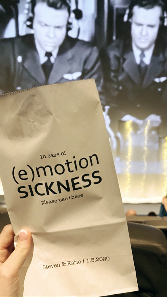 A paper bag reading “In case of (e)motion sickness please use these. Steven &amp; Katie | 1.5.2020” is held up in front of a theater screen playing a vintage airline travel advertisement in black and white. The screen shows two men in pilot uniforms wearing headsets.