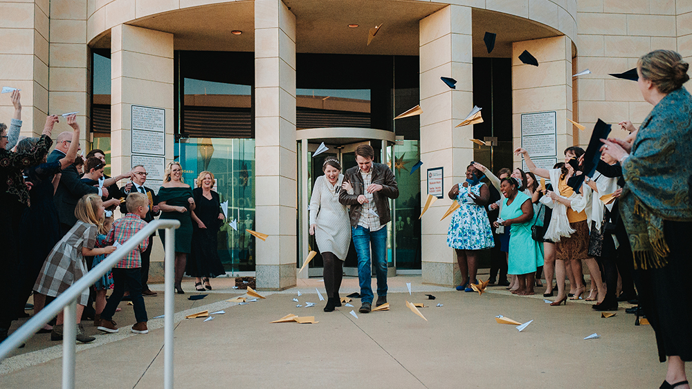 Katie and Steven, former aerospace engineering students, are dressed in casual wedding reception clothes. They walk out of their wedding venue with guests lined up along each side. Katie and Steven smile as they walk out to a shower of navy blue and gold paper airplanes being thrown by their guests.