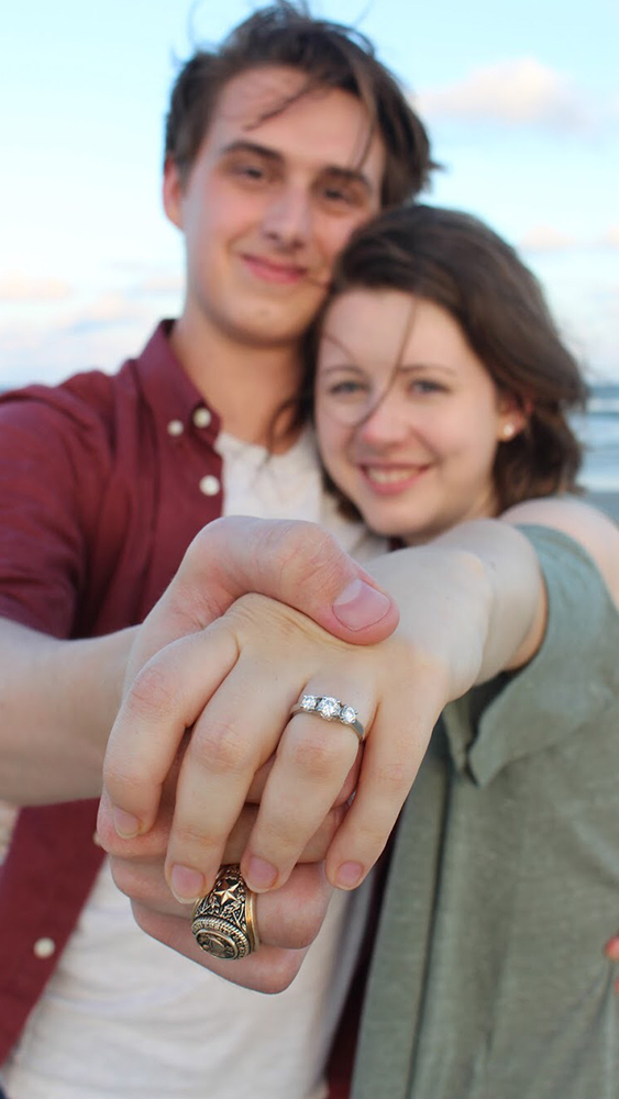 Katie and Steven, former aerospace engineering students, pose together on the beach immediately after their proposal. Steven is holding Katie’s hand out for the camera, showing her new engagement ring. Steven’s Aggie Ring is also in clear view.