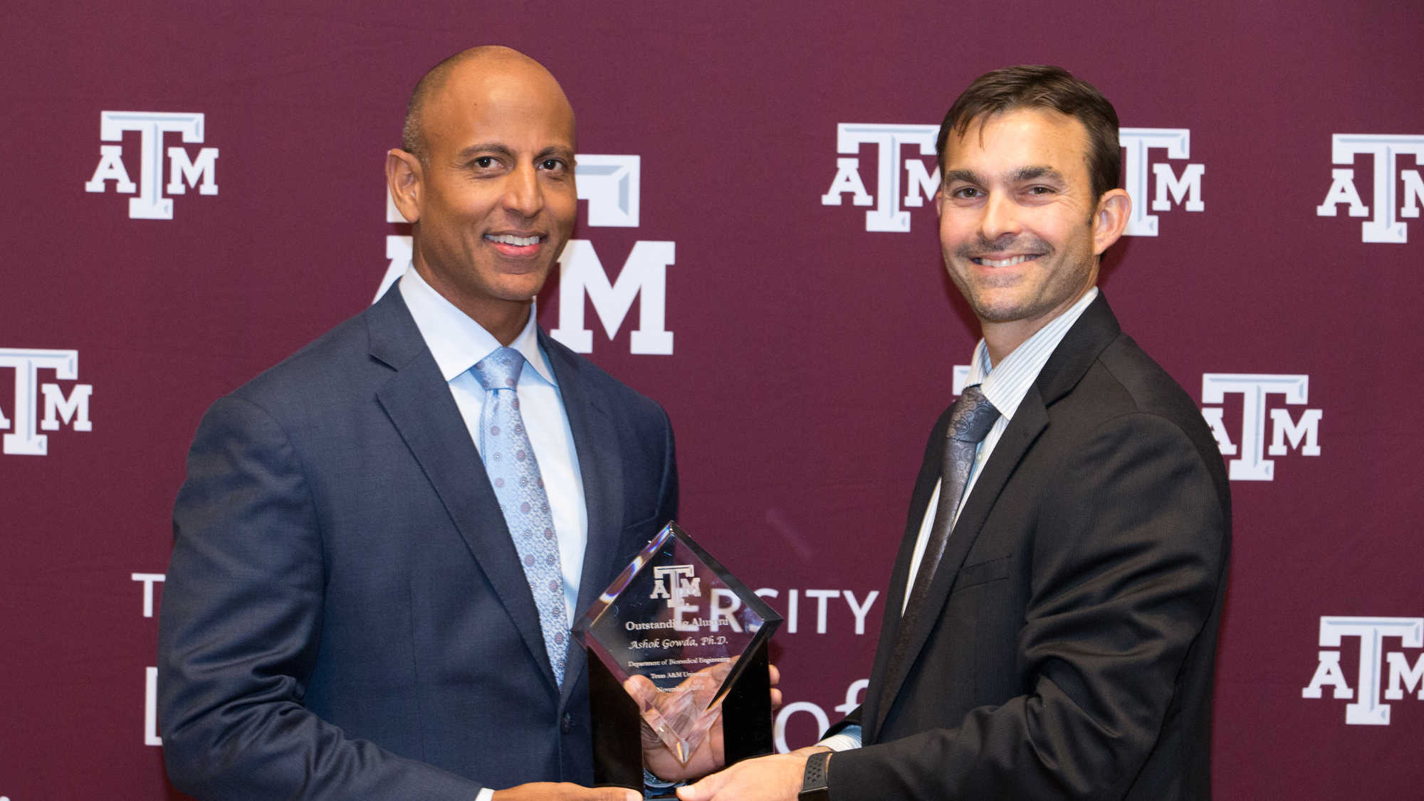 Two men in suits standing in front of maroon banner with white Department of Biomedical Engineering logo. They are holding a trophy made from clear crystal with the words "Outstanding Alumni, Ashok Gowda, Ph.D." etched on it.