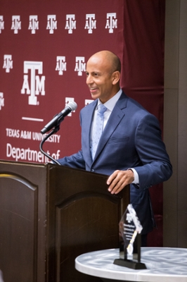 Man wearing a blue suit standing at podium smiling. Behind him is a maroon banner with the Department of Biomedical Engineering logo in white.