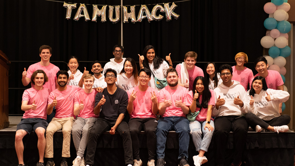 Smiling TAMUhack organization members sitting on a stage in front of a black backdrop (with balloon letters that spell out TAMUhack across the top) giving "gig 'em" hand sign. 