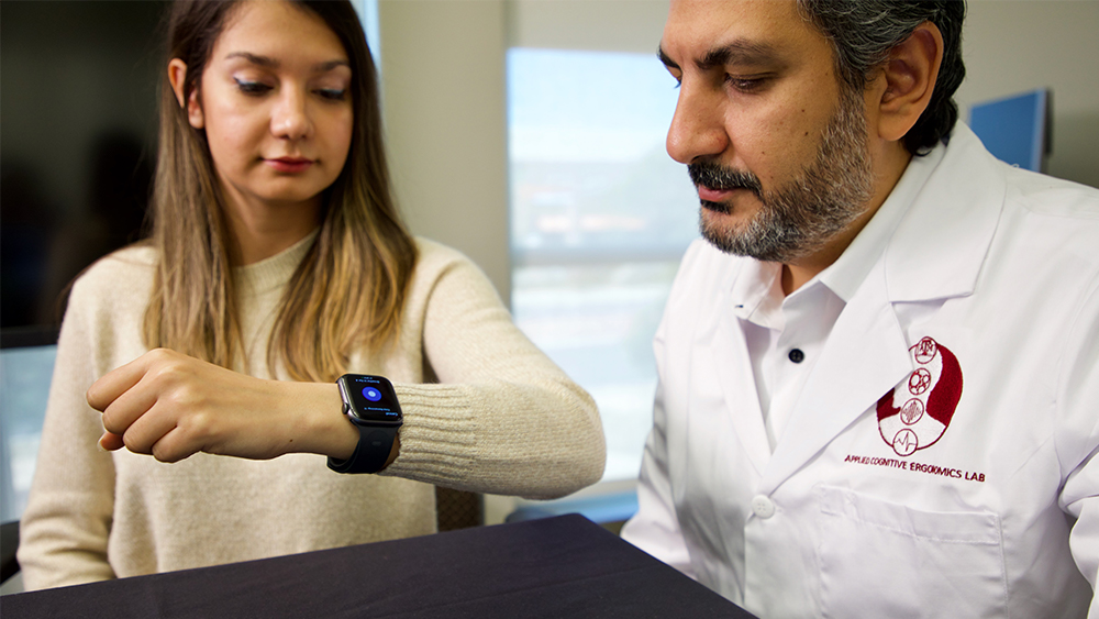 Student and professor in a laboratory both look at a smartwatch on the student's wrist. 