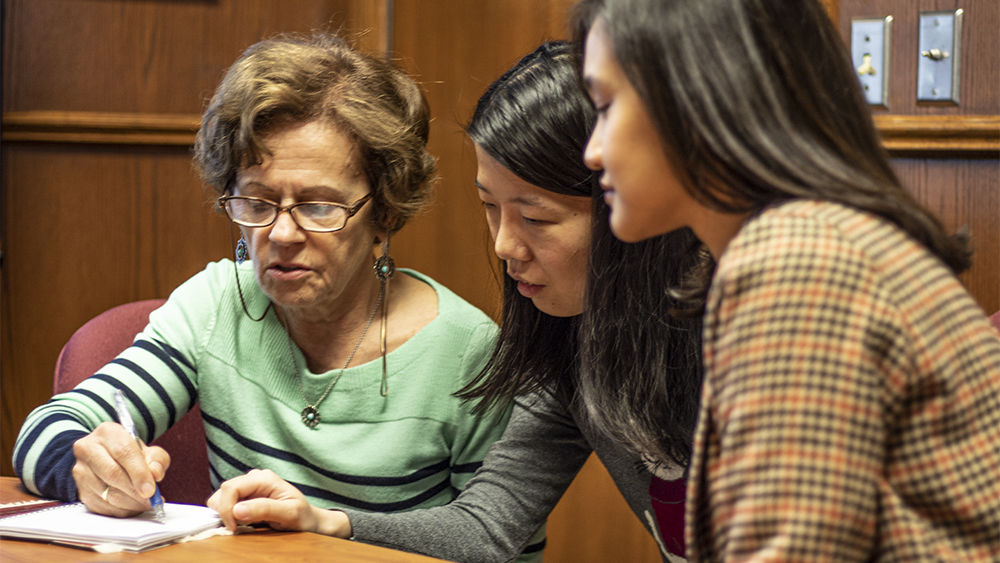 Female faculty member explains research notes to two female graduate students while all are seated at a table.