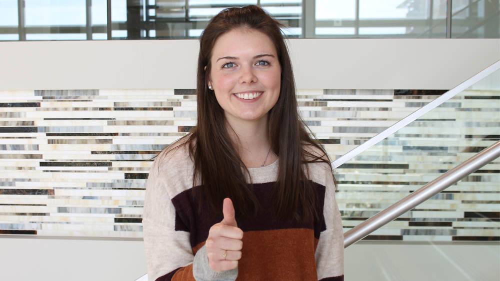 Photo of girl with brown hair and gray eyes standing on stairs smiling. She is giving the thumbs up, Gig 'em signal.