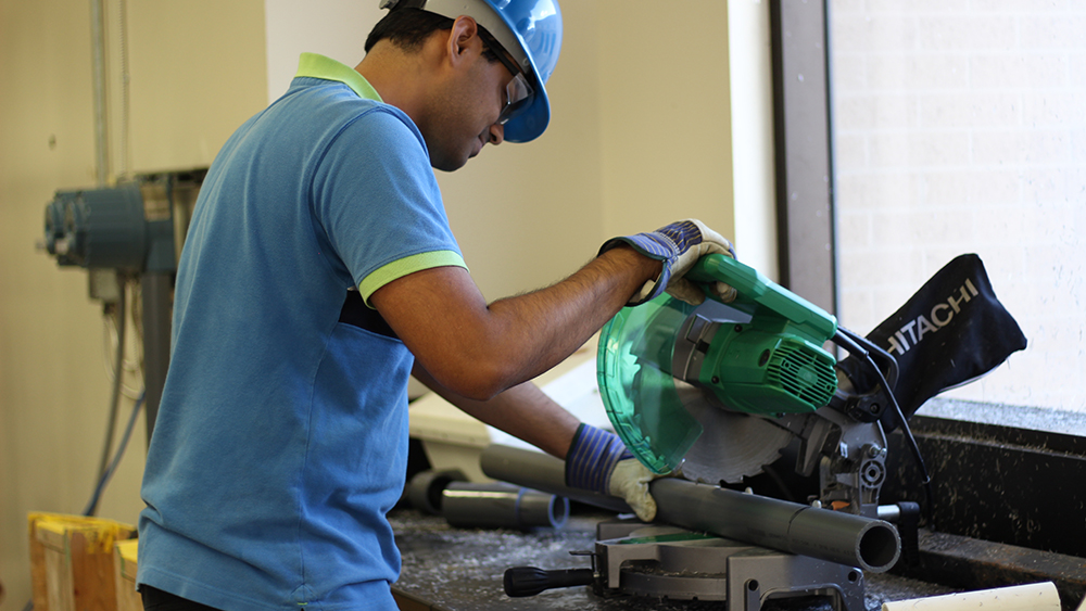 graduate petroleum engineering student Kaushik Manikonda wearing a hard hat and safety gear while cutting lengths of PVC pipe with a portable chop saw