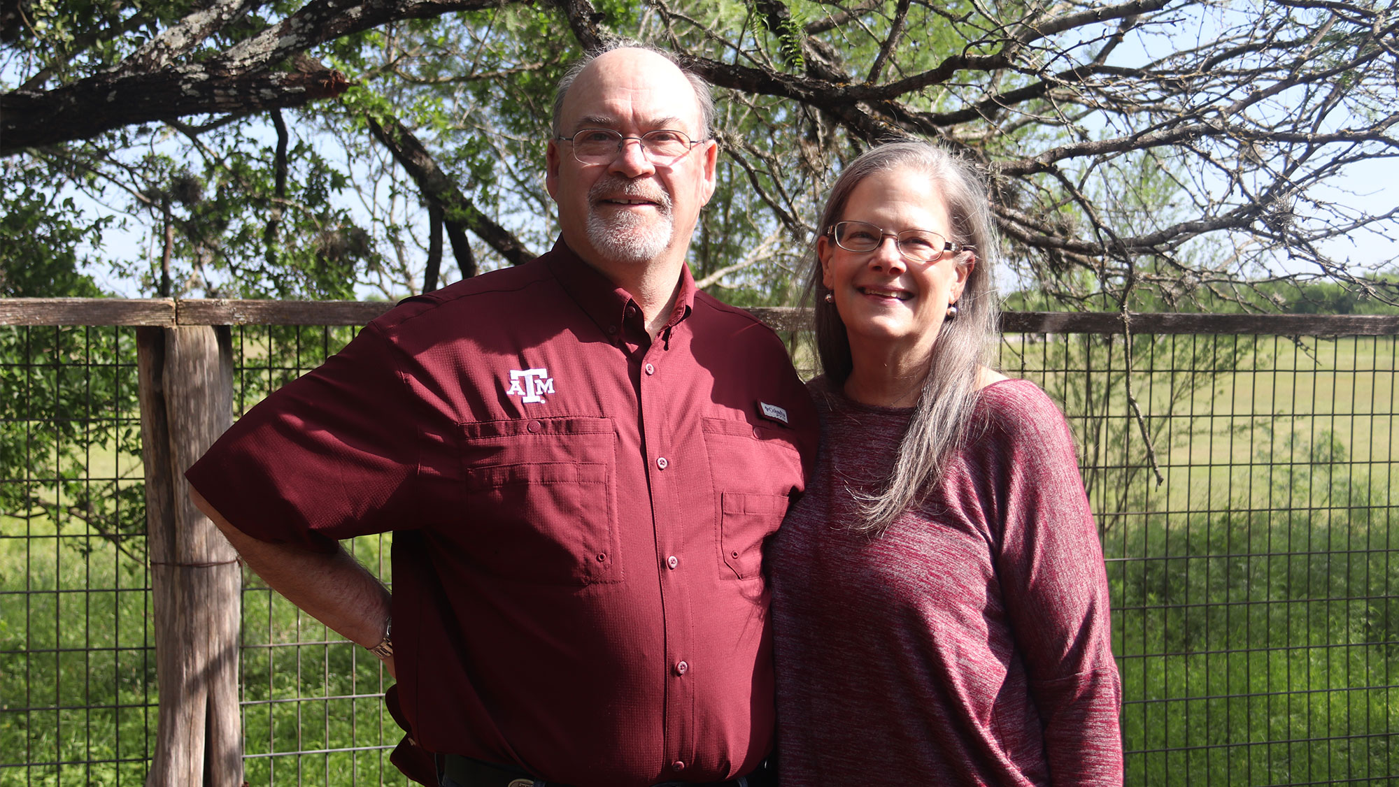 Keith and Lee Coleman stand outside in front of a tree and fenced pasture.