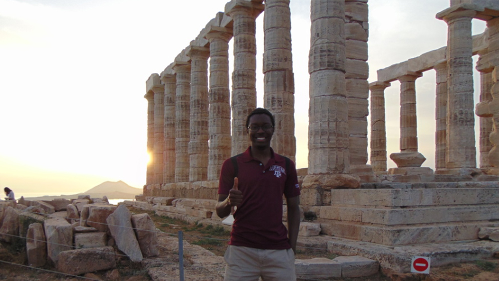 Student stands in front of Greek ruins. 