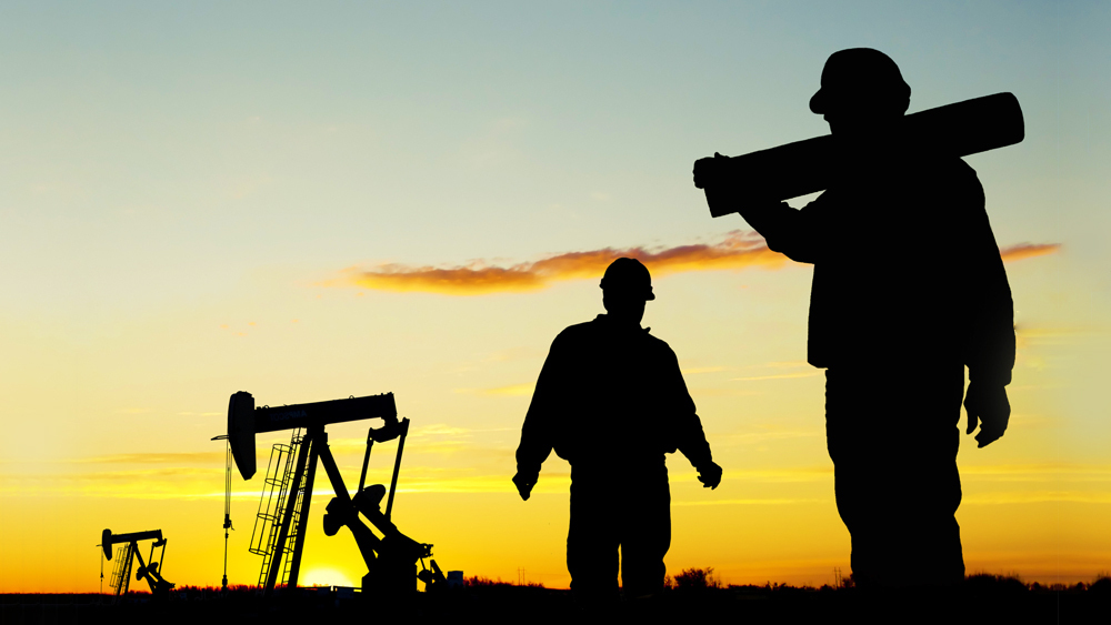 Two oil field workers at a well site with pump jacks in the background.