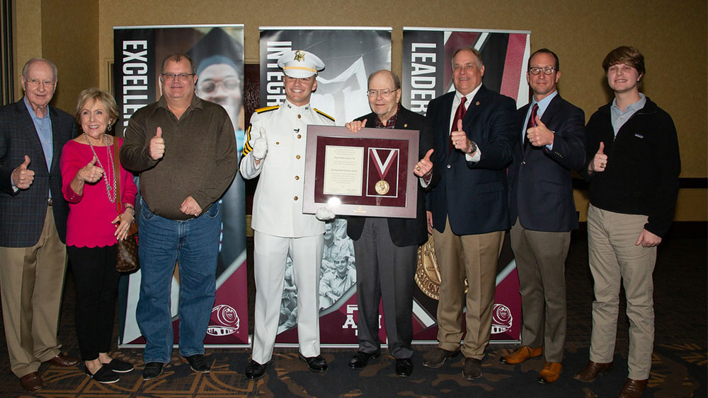 Weldon Jaynes receives a plaque detailing his achievements from a Ross Volunteer dressed in white, surrounded by Aggies doing a "gig'em" hand sign. 