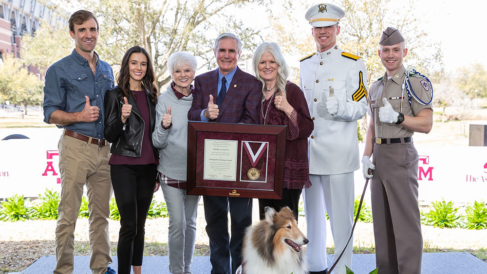 Tim Leach, surrounded by his family and a Ross Volunteer holds a plaque detailing his achievements. Reveille, the Texas A&amp;M mascot, sits at their feet.