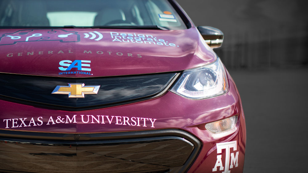 Front view of the Texas A&amp;M AutoDrive team's maroon autonomous car. Decals for General Motors and SAE International are on the hood of the car and there is a Texas A&amp;M University decal on the front bumper.