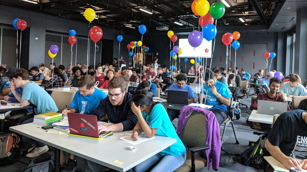 A large room filled with Texas A&M students sitting at tables in groups of two or three during a programming contest. On each table is a single, open laptop that the students are working on. There are multi-colored balloons tied to the backs of some of their chairs.