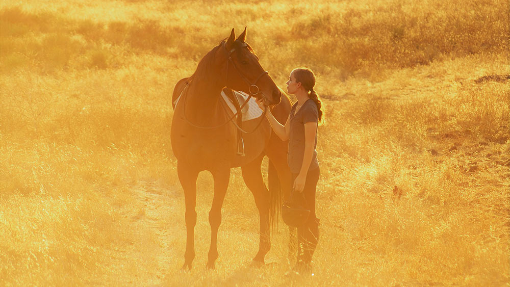 A woman stands in a field dressed in riding gear. She's standing next to her horse and looking into her eyes. 