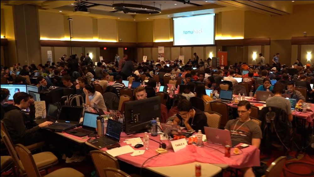 Students sitting at long tables in a large ballroom working on laptops and desktop computers during a hackathon competition.