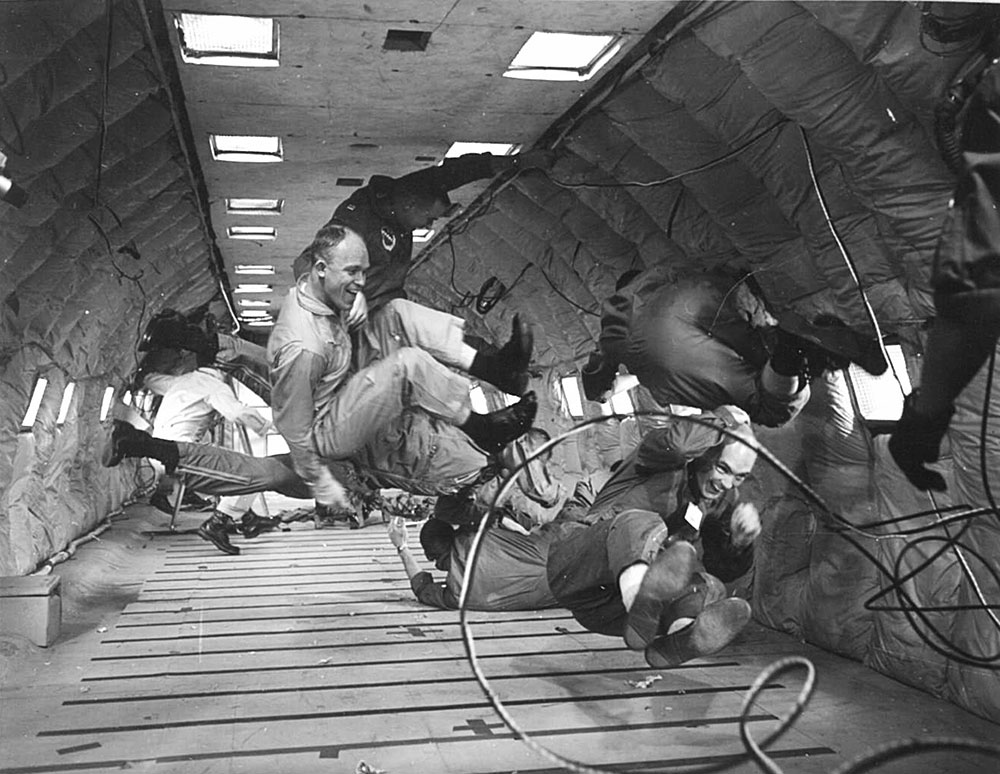 Black and white photo of men floating around the back of a padded airplane, testing zero gravity.
