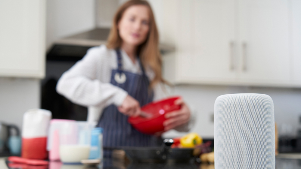 Woman wearing an apron preparing food in her kitchen. She is holding a red mixing bowl and is focused on an Alexa device on the counter in front of her.