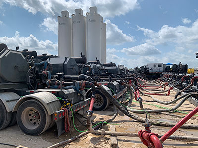 several tall tanks stand behind a long row of semi-trucks outfitted with pumping hoses for fracturing operations