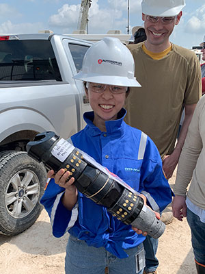 smiling female student wearing hard hat holds two-foot long cylindrical well plug for fracturing while male student wearing hard hat stands behind her and smiles 