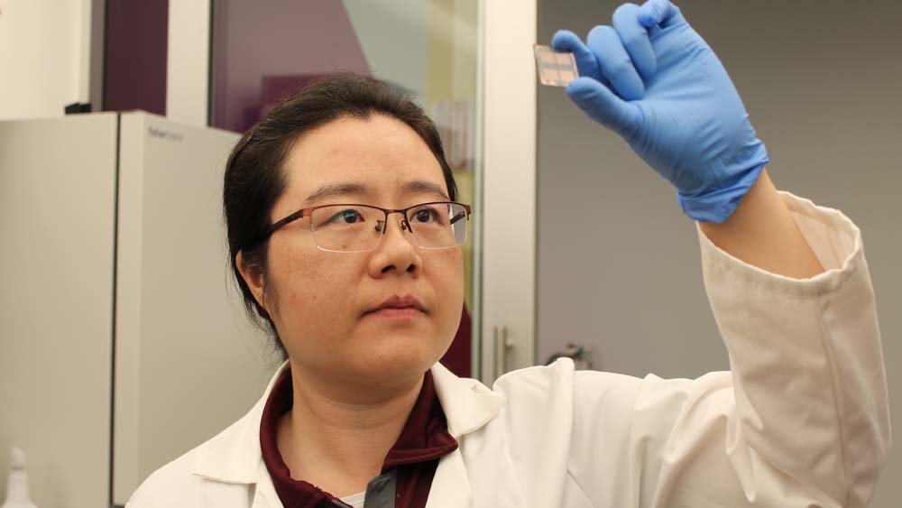 Female researcher in a lab holds a small square with a sensor on it up to the light. She is wearing gloves and glasses.