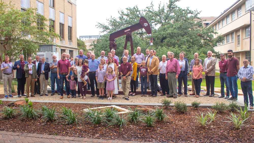 Crowd of nicely dressed people of various ages smile and pose for picture in front of pumpjack on Texas A&amp;M University campus
