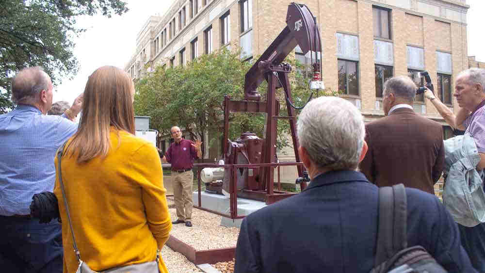 Man gestures to pumpjack behind him while speaking to a crowd of people on the Texas A&M University campus