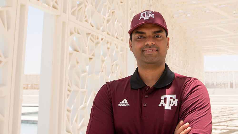 Male student wearing Aggie hat and shirt smiling while standing under white latticed structure on Texas A&M University of Qatar campus