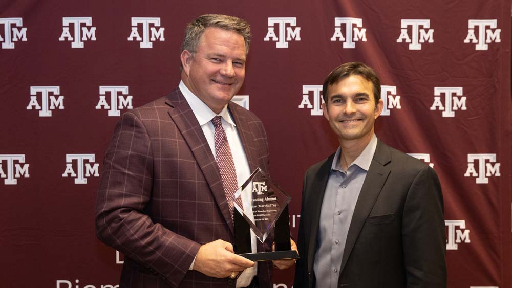 Dr. Winston Marshall and Dr. Mike McShane holding clear award plaque. Behind them is a maroon banner with small Texas A&amp;M University logos throughout.