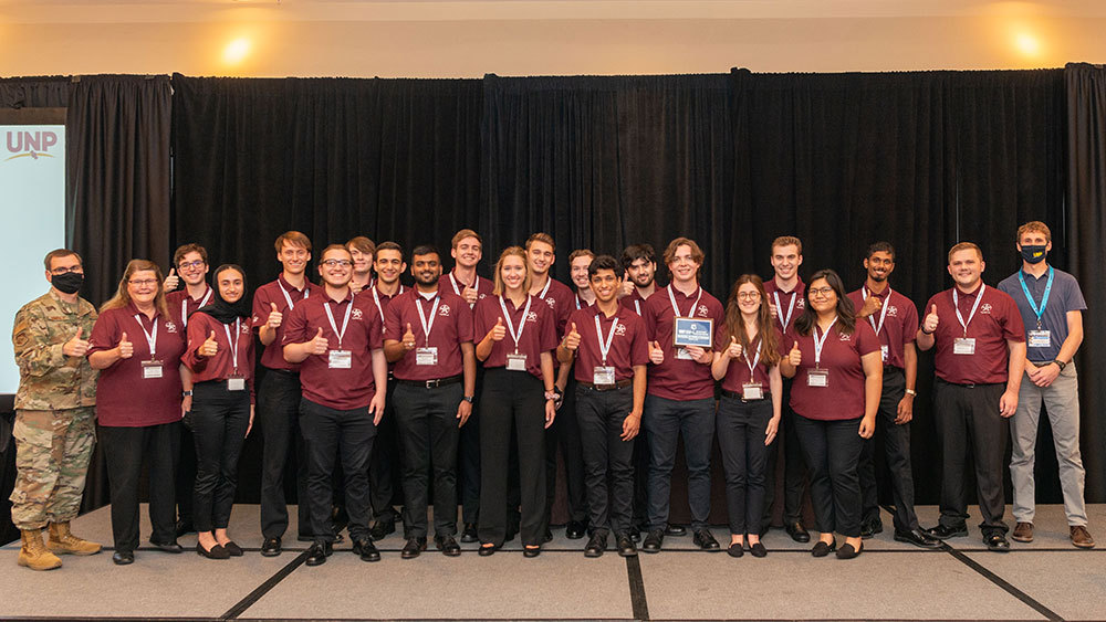 Texas A&M University students in maroon polos taking a group photo on a stage after receiving a University Nanosatellite Program winner plaque.