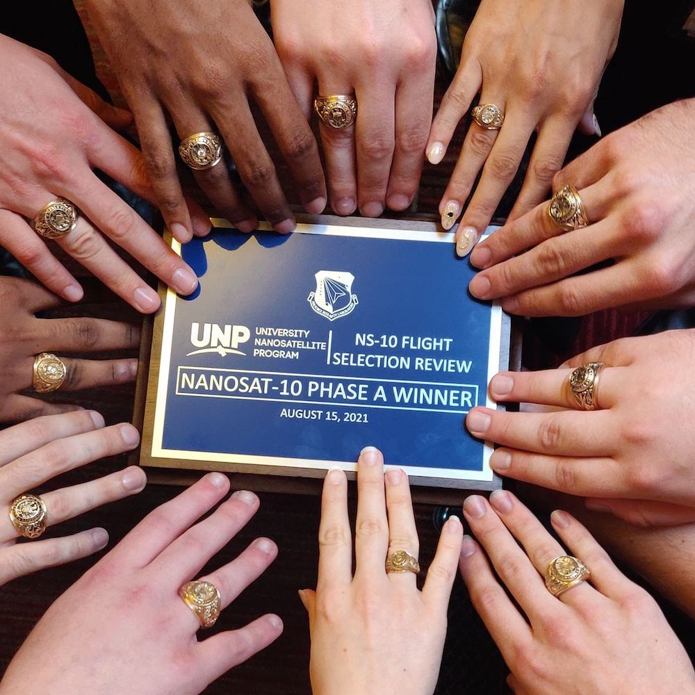 Texas A&amp;M University students' hands in a circle around a University Nanosatellite Program winner plaque, showing their Aggie rings.