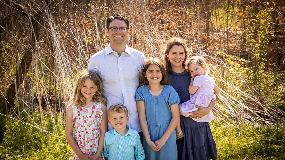 Mike Frizzell and his wife stand in a wooded area with their four children.