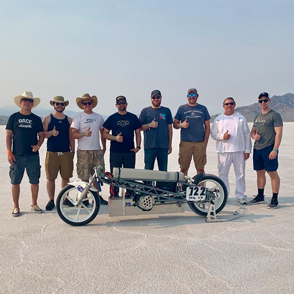 eight men stand together behind an electric motorcycle. the men are donned in jeans, cargo shorts and t shirts. they are wearing sunglasses, cowboy hats and giving the camera a thumb's up