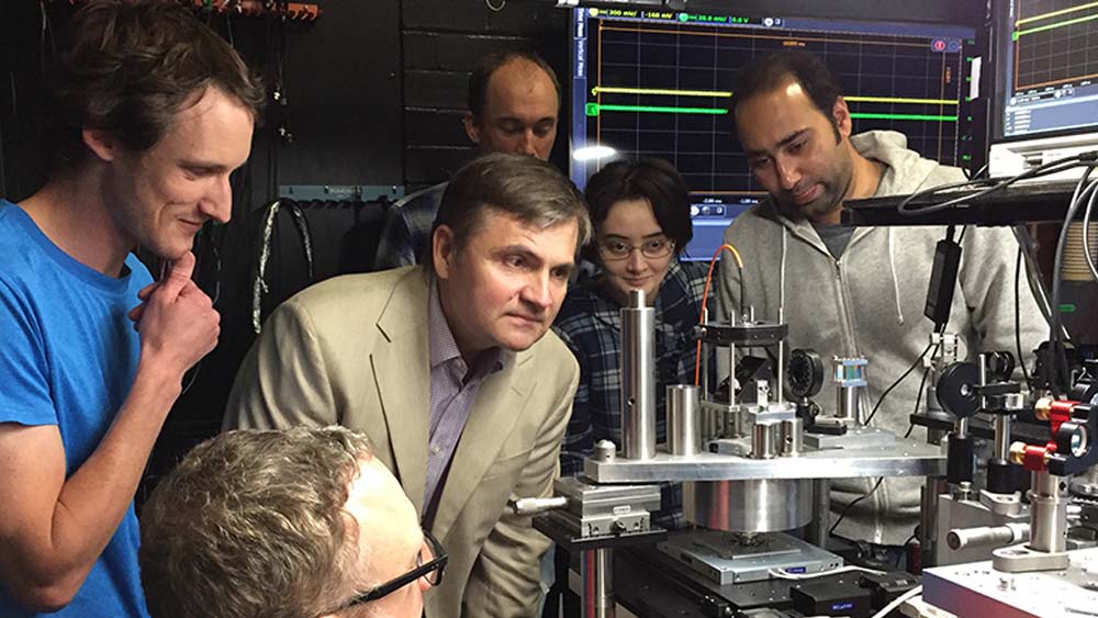 A professor and students stand gathered around a laser.