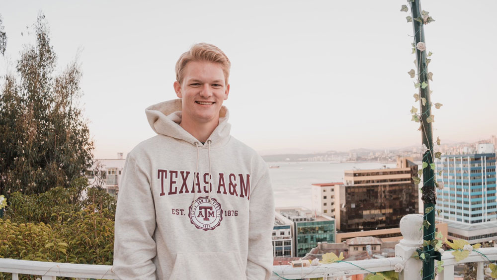 Tanner Hoke smiling at the camera. Behind him is a white rail with the view of a few city buildings directly behind him and a few more in the distance further back.