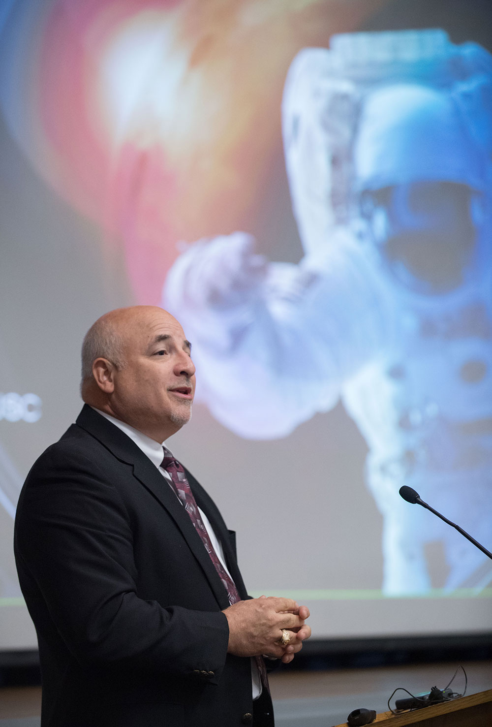 Joe Caram giving a NASA presentation at a podium with an astronaut image behind him.