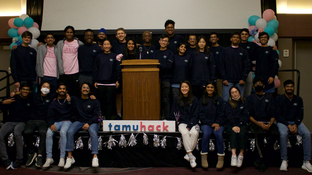 A group of students on a stage smiling at the camera. One half of the group are sitting on the edge of the stage and the other half are standing behind them. There is a podium in the middle of the stage with a sign that says "TAMUhack" in pink and blue letters in front of it.