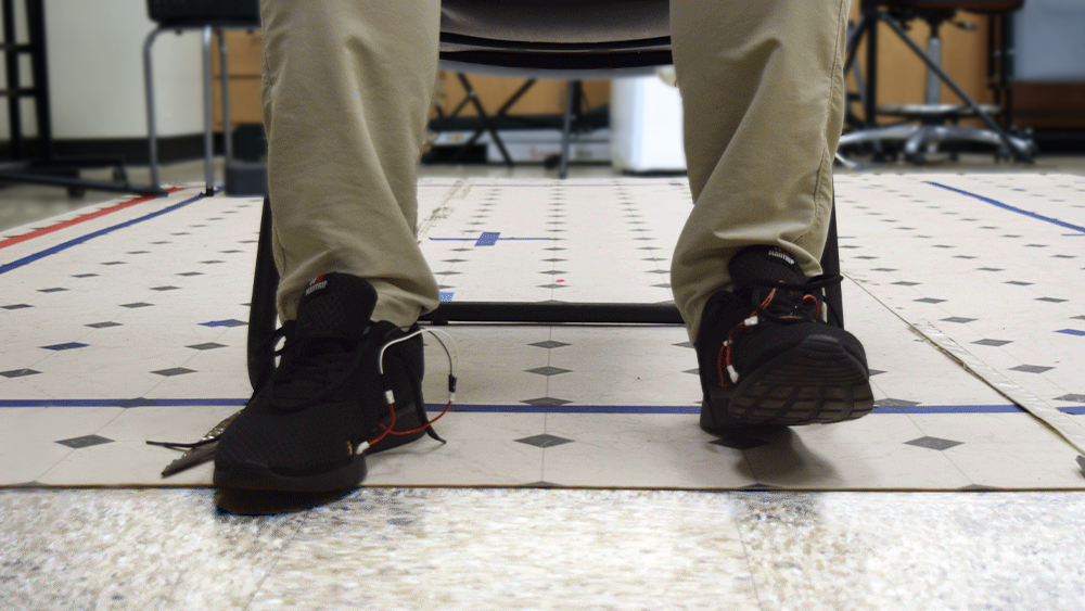 Seated person wearing shoes with sensors tapping their feet on the floor. 