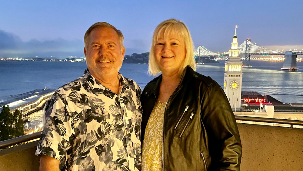 Claire and John Vassberg smiling together in front of the San Francisco-Oakland Bay Bridge.