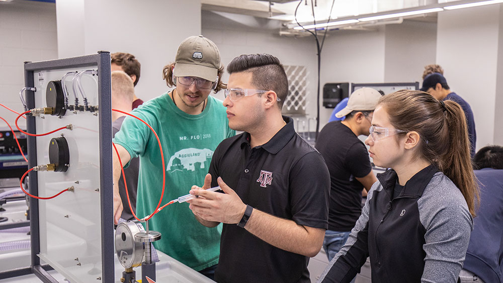 Three Texas A&M undergraduate students work with a syringe and tubes attached to a board in a laboratory among other students.
