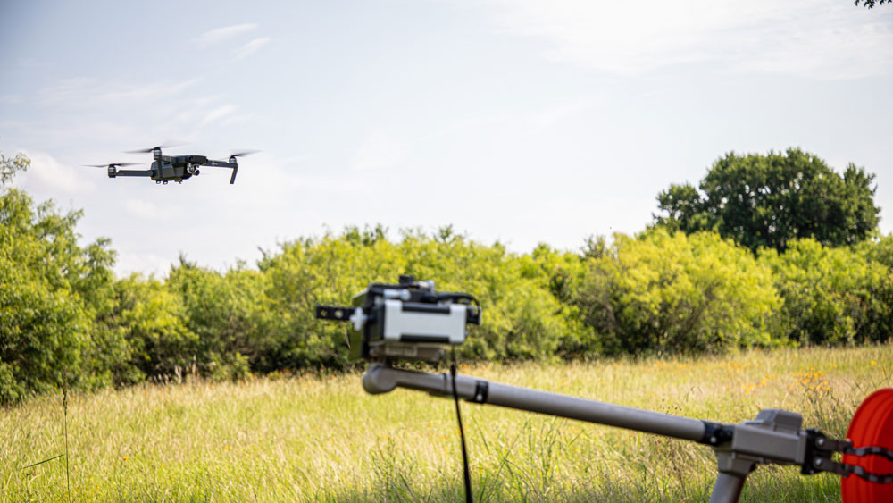 Aerial drone being flown in grassy field and tracked by camera mounted on rod.
