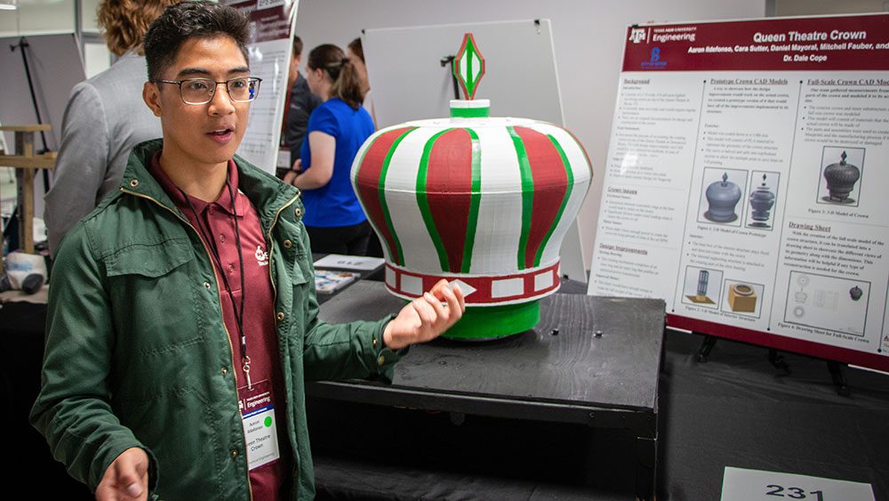 Male student at a Capstone project competition discusses the model of a rotating royal crown in front of a poster showing the crown’s interior mechanics
