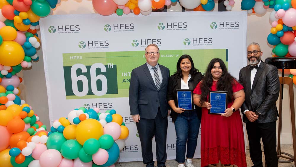 Dr. Ranjana Mehta and Oshin Tyagi hold plaques and stand between two men in front of a colorful balloon arch and a Human Factors and Ergonomics Society banner. 