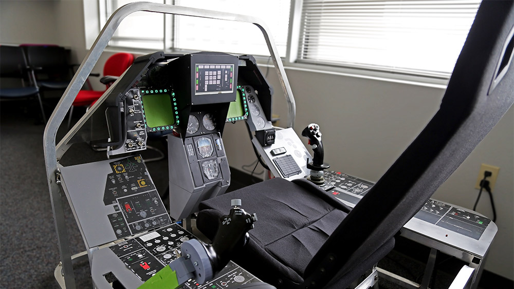 An aluminum-framed flight simulator equipped with hand on throttle and stick controls, a multifunctional display, toggle switches and stickers replicating the layout of an F16.